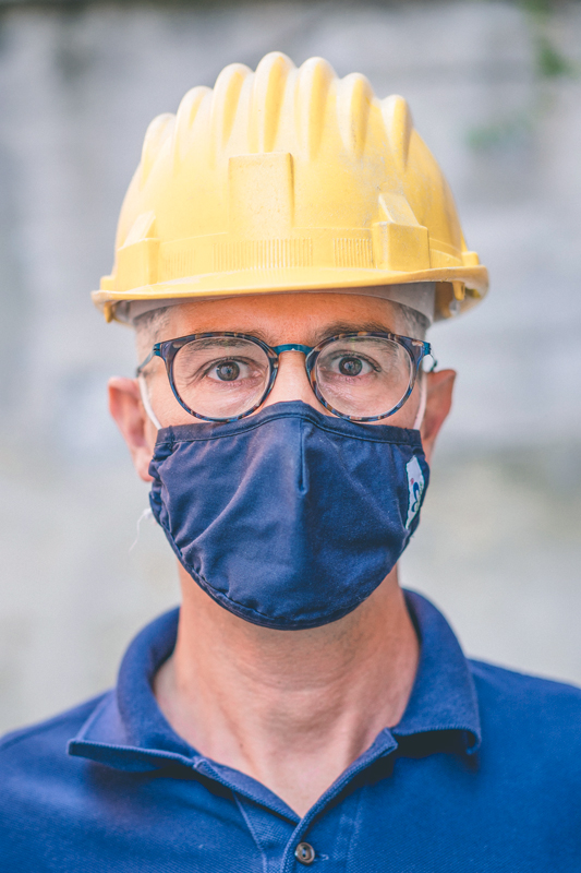 Alessandro is looking into the camera wearing a security helmet. He also wears a mask for covid protection.