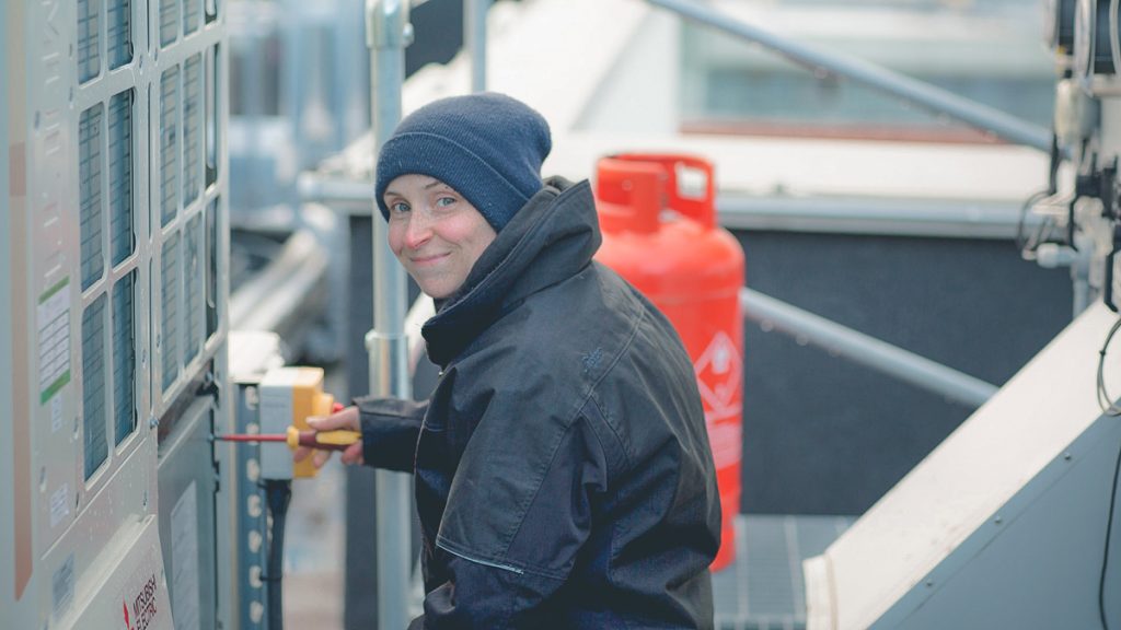 Ruth smiles at the camera while maintaining an HVAC unit.