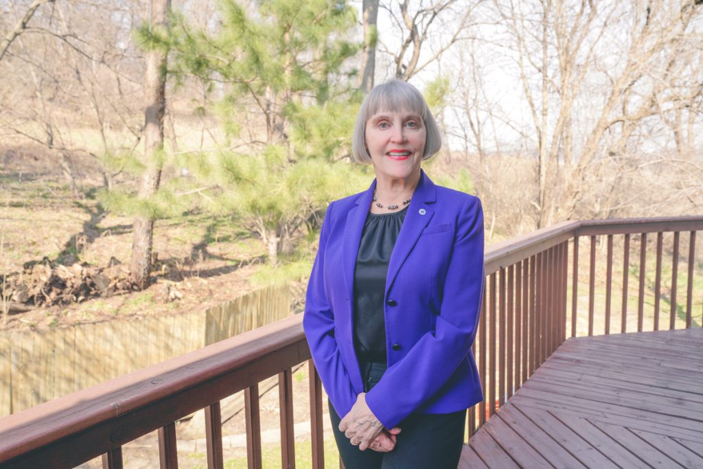 Renee smiles for the camera. She is on a porch. Behind her is a beautiful natural landscape.